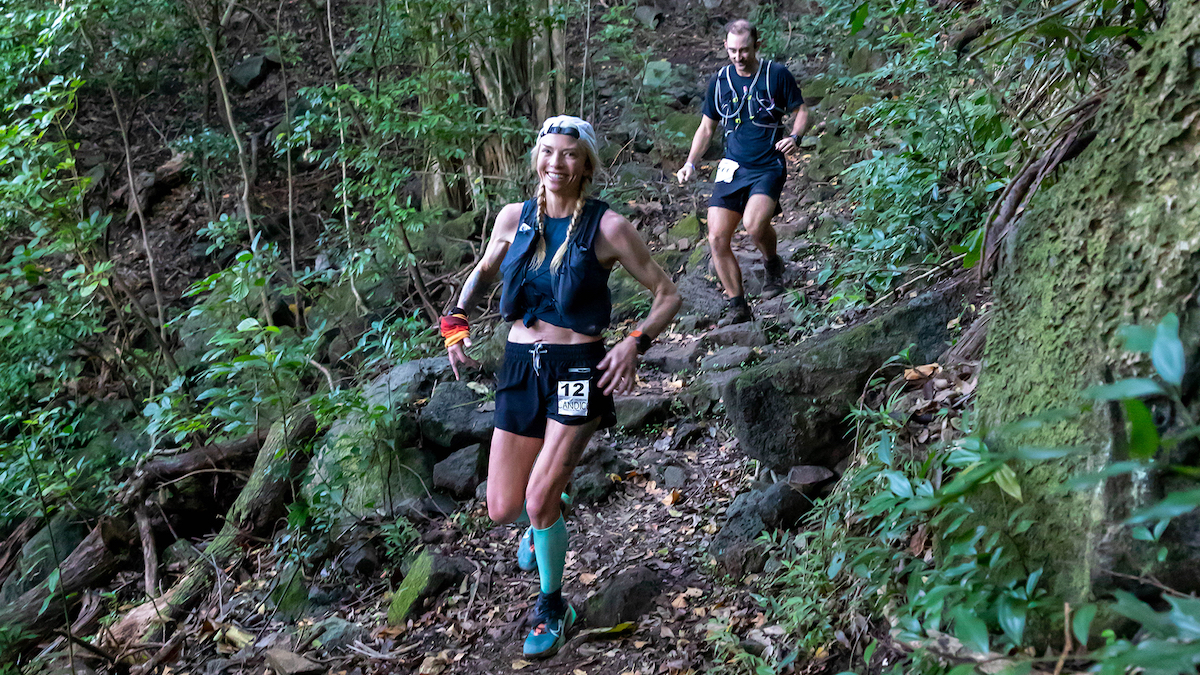 A woman and her pacer run in Hawaii on rooted trail. They are smiling and the forest is green.