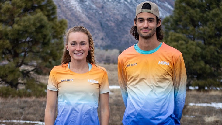 Two runners, one female-bodied, one male-bodied, stand with orange/blue shirts in front of a forested background