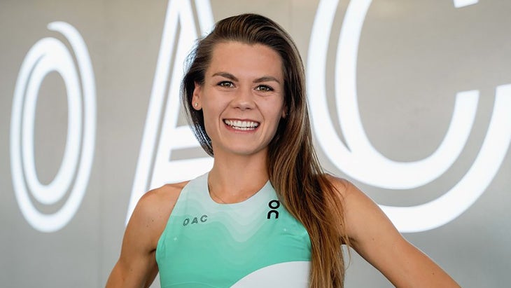 A female-bodied runner stands with brown hair and a green shirt, in front of a grey backdrop with white lettering