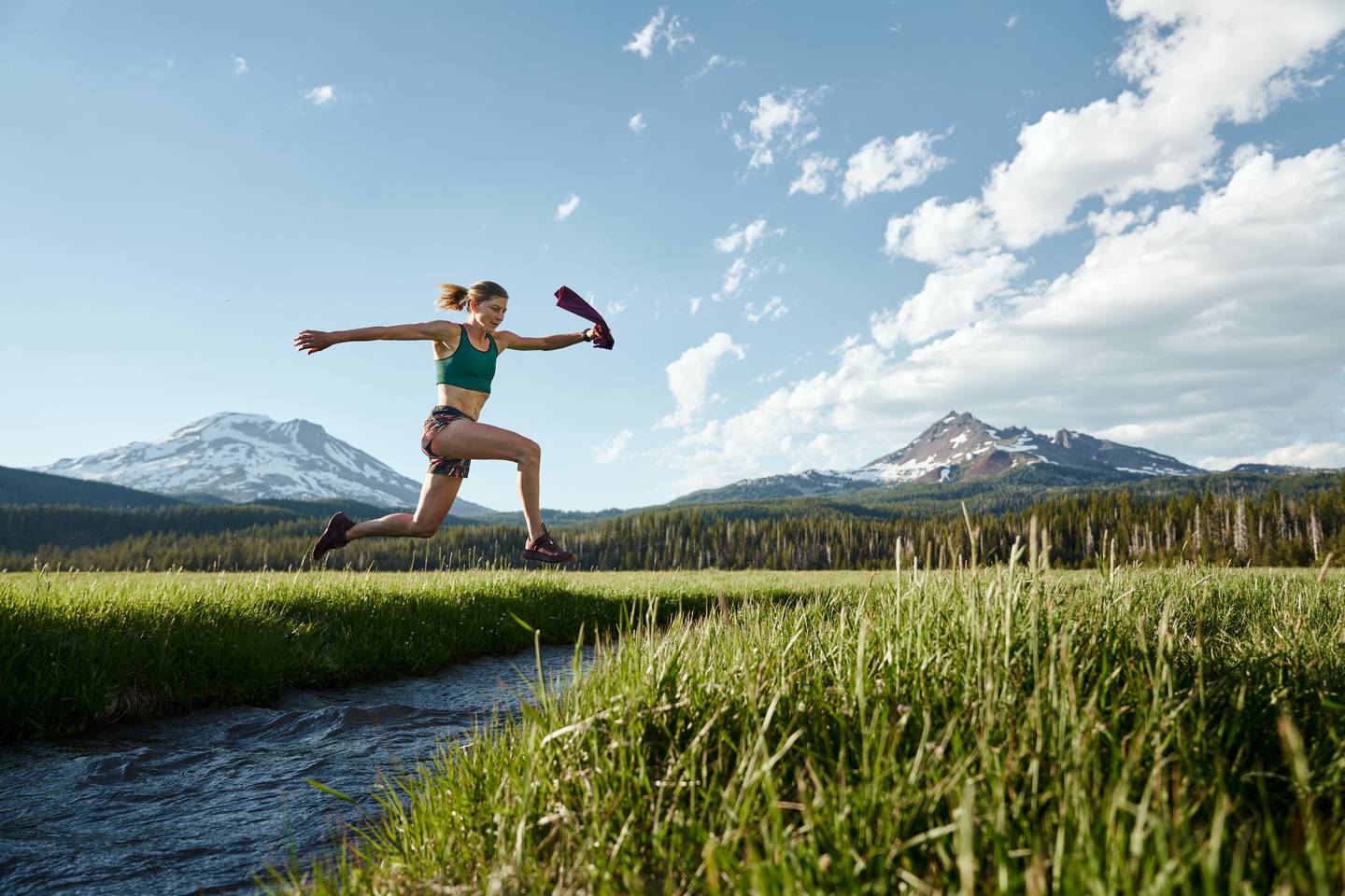 Lauren Fleshman leaps across a waterway with a mountain scene behind her
