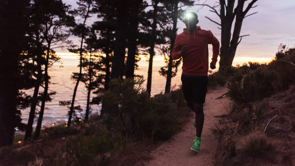 Focused young African man wearing a headlamp running alone down a trail in the forest while out for a cross country run at dusk.